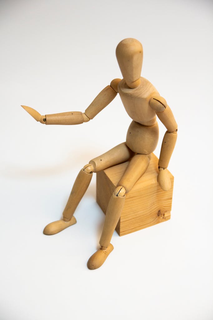 A wooden artist's mannequin sitting on a wooden block with one arm raised, set against a white background.